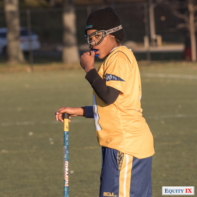 Girl with black hat and goggles and mouthguard wearing yellow jersey and lacrosse stick in NYC representing wealth disparity issue for sports © Equity IX - SportsOgram - Leigh Ernst Friestedt