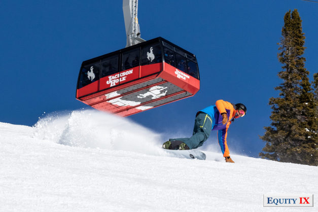 Matt Friestedt turns on snowboard with red Jackson Hole tram behind him on mountain with snow spraying from the edges of the snowboard © Equity IX - SportsOgram - Leigh Ernst Friestedt